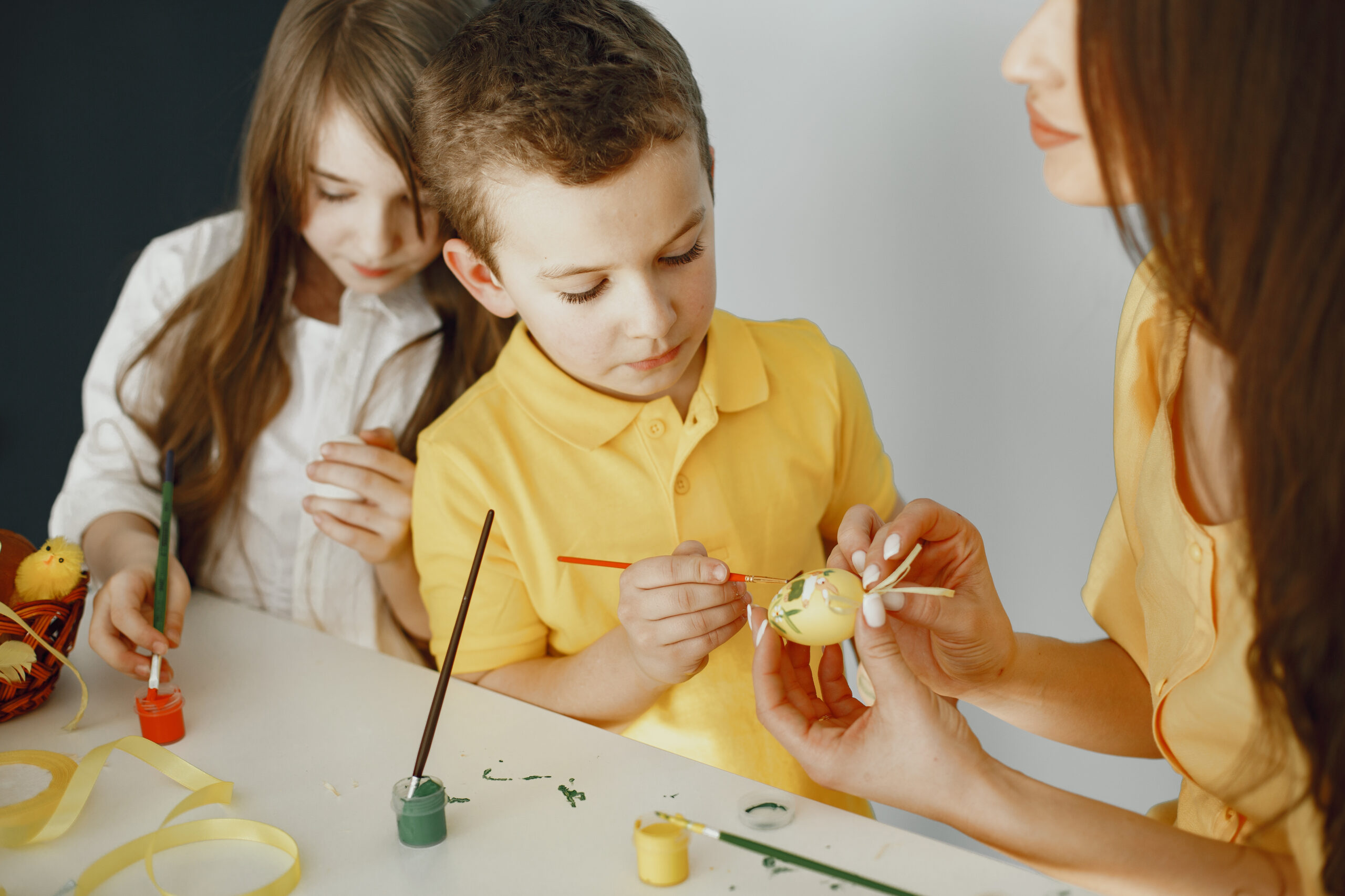 Mom teaches children to draw Easter eggs at table in yellow clot
